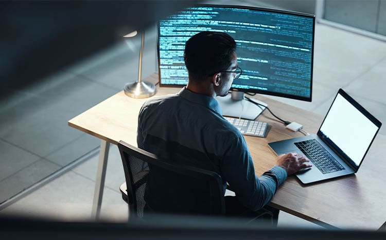 Professional software developer working with dual monitors displaying programming code and laptop computer system