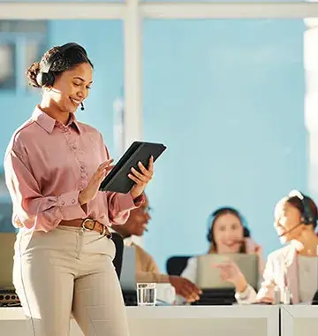 Smiling customer support agent wearing headset using tablet device with team working in bright modern call center